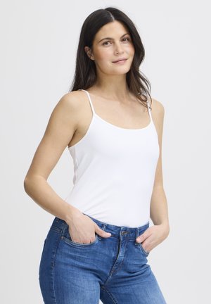 Woman with long dark hair wearing a white spaghetti strap top and blue jeans, hands in pockets, against a plain light background.
