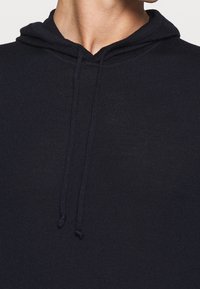 Marinblå huvtröja med en texturerad tygkvalitet, synliga snoddaccent och en figurnära design runt halsen och axlarna.