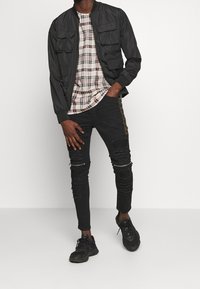 Chaqueta bomber negra con dos bolsillos frontales, combinada con una camiseta a cuadros y jeans negros rasgados, que cuentan con una raya lateral, y zapatillas negras.