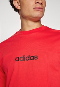 Raudona medvilninė marškinė su juodu "adidas" logotipu priekyje. Medžiaga atrodo lygi, su apvaliu kaklu.