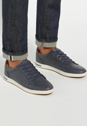 Blue leather sneakers feature a textured side panel, white sole, and round toe, paired with dark denim pants rolled at the cuffs.