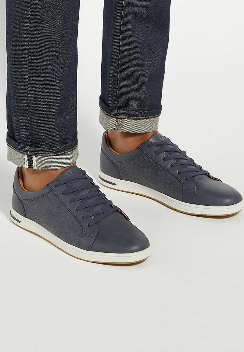 Blue leather sneakers feature a textured side panel, white sole, and round toe, paired with dark denim pants rolled at the cuffs.