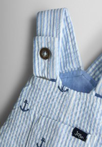 Salopetă cu dungi albastre deschise și albe, cu o bretea cu nasturi pe umăr, ancore brodate și o etichetă mică de marcă pe buzunar.