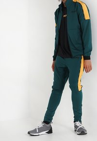 Grön och gul träningsoverall med zip-jacka och byxor, ihopparad med grå sneakers. Har varumärkeslogotypen på toppen och sidodetaljer.