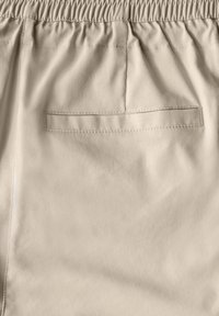 Pantaloni beige corti con cintura elastica, dotati di una tasca posteriore con patta. Texture liscia con un design strutturato, dettagli di cucitura minimi.