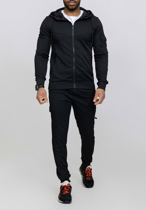 Survêtement de sport noir avec une veste à capuche zippée et un pantalon de style cargo. Présente des poignets côtelés et deux poches latérales. Les chaussures ont des accents orange.