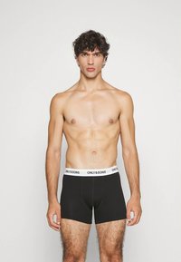 Mustad bokserid valge elastse vöökohaga, millel on "ONLY&SONS" logo. Lühikeste lokkis juustega mees seisab ühevärvilise tausta ees.