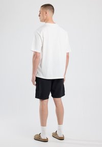 White cotton t-shirt with short sleeves, relaxed fit. Paired with black shorts. Displaying a minimalistic design with a back view.
