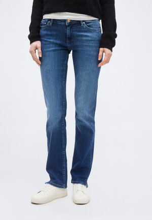 Άτομο που φοράει μπλε τζιν ίσιας γραμμής, λευκά αθλητικά παπούτσια, μαύρο πουλόβερ και λευκό πουκάμισο ορατό στη μέση σε απλό φόντο.