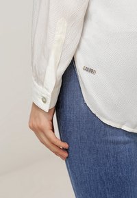 Ruka odpočívající podél těla, oblečená v bílé texturované košili s dlouhým rukávem a knoflíkovými manžetami, a v modrých džínách.