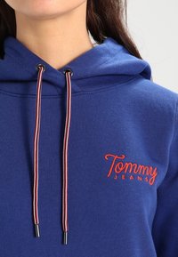 Felpa blu con cappuccio, dotata di logo ricamato rosso "Tommy Jeans", e lacci con un motivo rosso e bianco e punte in metallo.