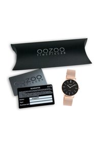 Montre noire avec des accents dorés et un bracelet en maille, présentée dans une boîte noire de protection et avec une carte de garantie.