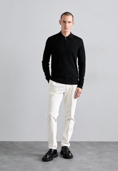 Sort langærmet polo med lynlås, hvide skræddersyede bukser og skinnende sorte loafers, stående mod en lysegrå baggrund.