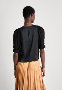Blouse en satin noir avec des manches bouffantes, col rond et détail de fermeture éclair au dos, associée à une jupe plissée beige. Conception simple et élégante.
