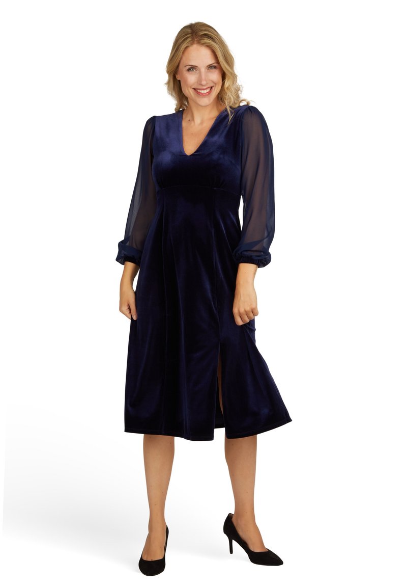 Navyblaues Samtkleid mit transparenten Puffärmeln, V-Ausschnitt und Frontschlitzen. Der Rock fließt bis zur Wade und wird mit schwarzen High Heels kombiniert.