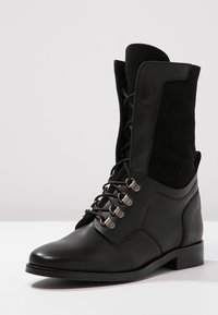 Botín negro con cordones hasta el tobillo, fabricado en piel y ante. Presenta un acabado liso, puntera redondeada y ojales metálicos para los cordones.