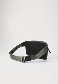 Melnas auduma jostas soma ar teksturētu virsmu, ar rāvējslēdzēju un regulējamu olīvzaļas krāsas siksnu, kurai ir Calvin Klein logs.