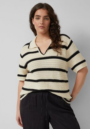 Femme aux cheveux bouclés portant un polo en maille à manches courtes rayé crème et noir et un pantalon noir ample sur un fond gris.