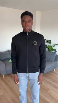 Νέος άνδρας που φορά μαύρο μπουφάν με φερμουάρ και μικρό λογότυπο στο στήθος και ανοιχτόχρωμα μπλε τζιν, στέκεται σε σαλόνι με καναπέ και φυτό.