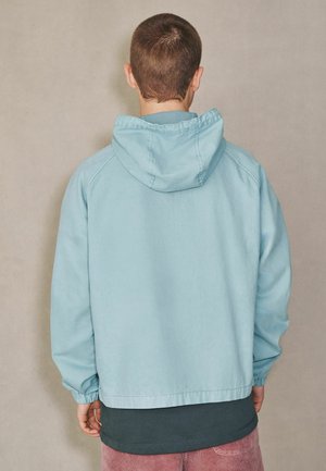 Person, die mit dem Rücken zur Kamera steht, trägt eine hellblaue Kapuzenjacke über einem dunklen Hemd und rosa Hosen vor einem schlichten beigefarbenen Hintergrund.