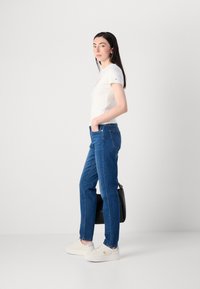 Donna in piedi con le mani nelle tasche dei jeans blu, indossa una maglietta grafica bianca, sneaker bianche e tiene una borsa nera.