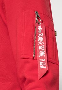 Rode trui met een zak met ritssluiting, voorzien van een contrasterend label met de tekst "REMOVE BEFORE FLIGHT". Zachte textuur en geribbelde details aan de manchetten.