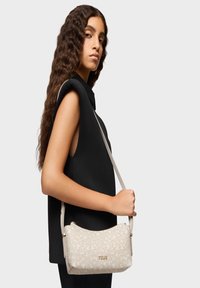 Bolso color beige claro con un patrón floral texturizado y un logo dorado. El bolso presenta una forma curva y una correa larga y ajustable.