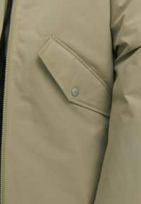 Section d'une doudoune beige avec une poche inclinée visible et un bouton-pression argenté. Texture lisse et mate, avec une fermeture éclair.