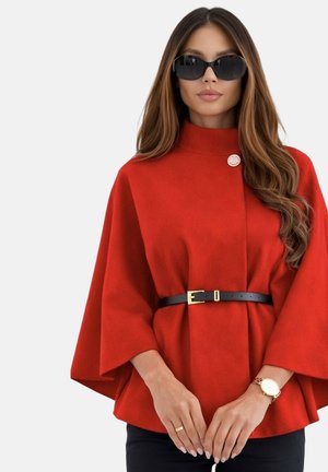 Femme portant des lunettes de soleil noires, un manteau cape rouge ceinturé, une montre en or et une bague, debout devant un fond blanc uni.