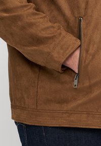 Veste en daim marron avec une poche zippée, texture douce et coupe ajustée, avec des coutures visibles le long de l'ourlet et des poignets.