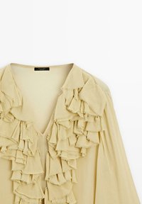 Beige bluse med V-hals og mange lag af flæser foran. Blødt stof med en let glans og uden opsynede kanter.