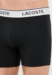 Fekete pamut boxer alsónadrág fehér derékkal, amelyen a "Lacoste" felirat szerepel. Jellemzője a szoros illeszkedés és a sima textúra.