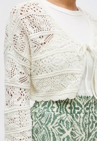 Λευκό πλεκτό ζακέτα με μοτίβα βελονιάς, δεμένο μπροστά, φορεμένο πάνω από λευκή μπλούζα και συνδυασμένο με ψηλόμεσο παντελόνι σε πράσινο χρώμα με σχέδια.