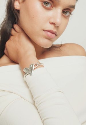 Jeune femme à la peau claire et aux yeux verts portant un haut blanc décolleté et un bracelet argenté en forme de cœur à son poignet.