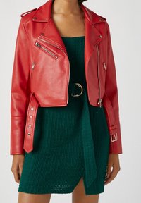 Röd läderkorthållig motorcykeljacka med dragkedjor och silverfärgad hårdvara, buren över en grön strukturerad stickad klänning med en sidoklyvning och bältesdetalj.