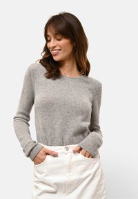 Kobieta uśmiechająca się i patrząca w dół, ubrana w szary sweter dzianinowy i białe spodnie, z rękami w kieszeniach, na jasnoszarym tle.