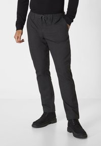 Schwarze Hose aus strukturiertem Stoff, mit elastischem Bund, verstellbarem Kordelzug und Seitentaschen. Getragen mit schwarzen Knöchelstiefeln.