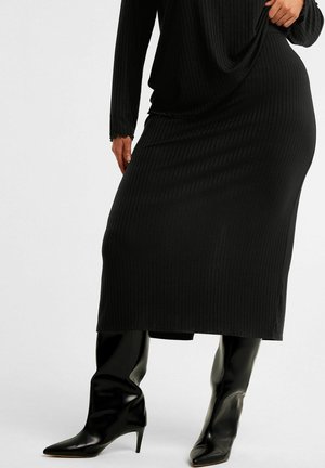 Robe noire côtelée à manches longues, avec une coupe ample et une longueur mi-mollet. Associée à des bottes pointues brillantes jusqu'aux genoux.