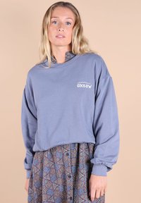 Sweatshirt gris oversize avec des poignets côtelés et un logo, superposé sur une jupe à motifs géométriques en bleu et marron.