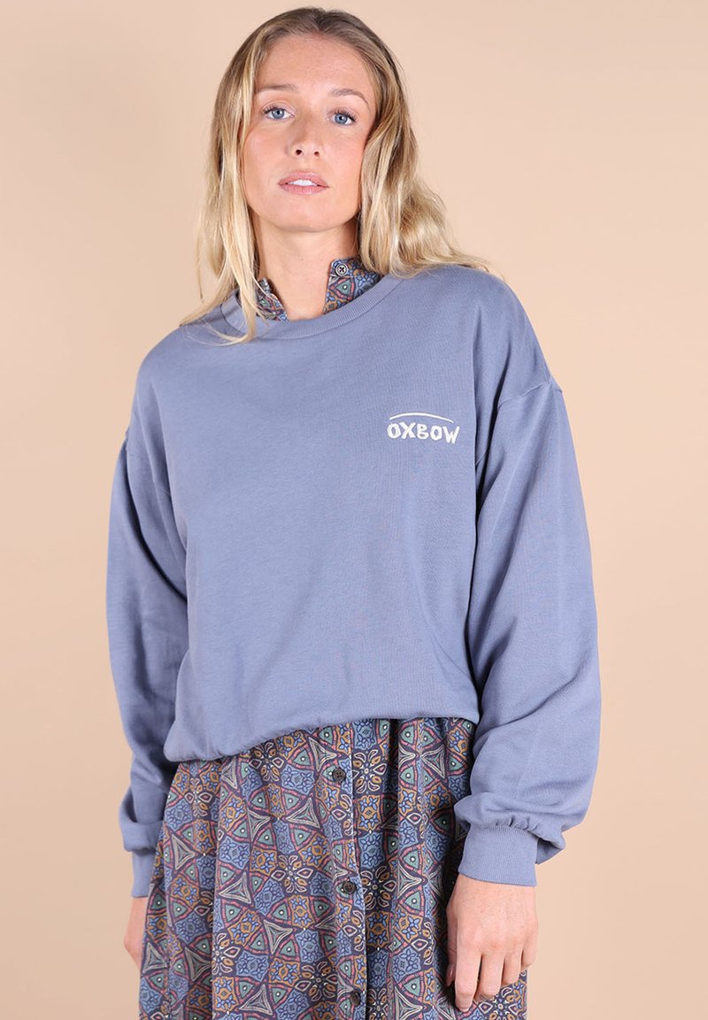 Sweatshirt gris oversize avec des poignets côtelés et un logo, superposé sur une jupe à motifs géométriques en bleu et marron.