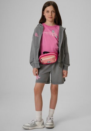 Młoda dziewczyna ubrana w szarą bluzę z kapturem i szorty z różowymi kwiatowymi wzorami, różowy T-shirt, różową saszetkę na biodra, białe skarpetki i białe trampki, stojąca na szarym tle.