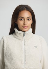 Fleeceväst i krämfärg med hög krage, textilstruktur, framsidan med dragkedja och logodetalj. Slät, figursydd innerskikt under.