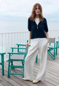 Vrouw met los bruin haar, gekleed in een zwarte gebreide cardigan, wit shirt, wijde witte broek en sneakers, staand op een houten dek aan zee.