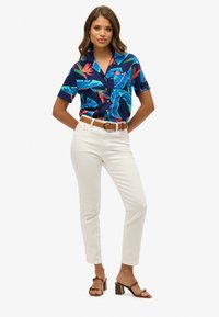 Korte mouwen, marineblauw shirt met een levendig bloemenpatroon; gecombineerd met nauwsluitende witte broek en bruine sandalen met hak. Voorzien van een borstzak.