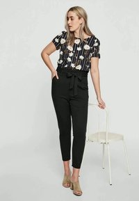 Floral patterned, short-sleeve blouse with navy stripes; high-waisted black pants with a tied belt; model standing beside a chair.