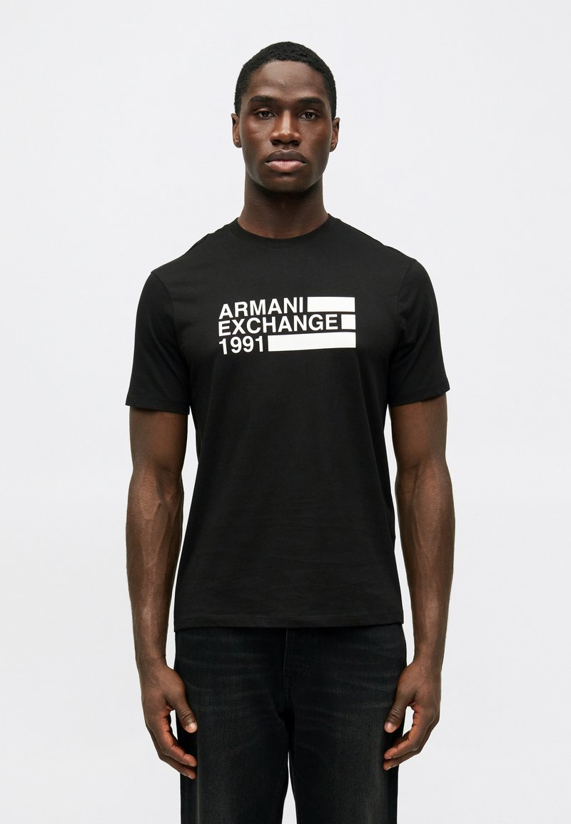 Άνδρας που φοράει μαύρο T-shirt Armani Exchange με λευκή επιγραφή "Armani Exchange 1991", στέκεται μπροστά σε λευκό φόντο.