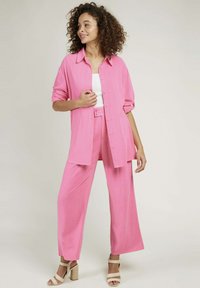 Chemise rose, ample, avec des boutons et des manches retroussées, assortie à un pantalon large. Le mannequin porte également des sandales à talons beiges.