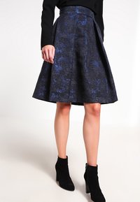 En knälång, utflytande kjol i mörk tyg med ett blått strukturerat mönster. Midjan är platt, och den är kombinerad med svarta ankelstövlar.