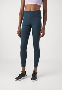 Leggings bleu foncé en matériau extensible, dotés d'une taille haute et d'une texture lisse. Associés à des chaussures de sport noires.