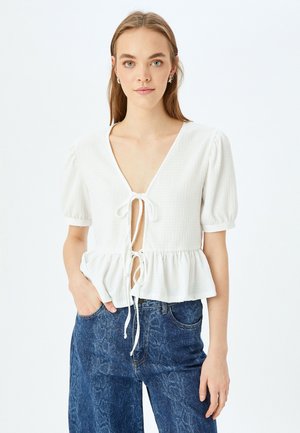 Blusa bianca texturizzata con maniche corte a palloncino, scollatura a V e chiusura con lacci sul davanti. L'orlo a peplum aggiunge volume e forma al design.